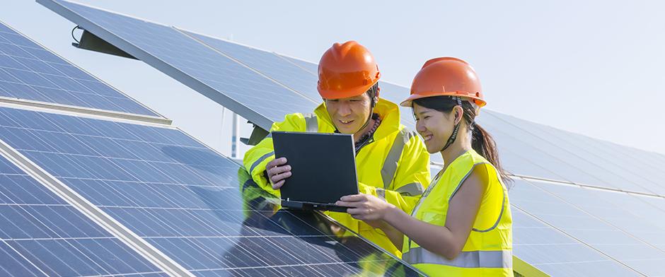 一男一女审视着笔记本电脑屏幕，旁边是一组工业规模的太阳能电池板.